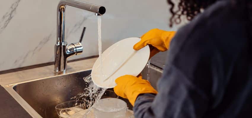 Person wearing yellow gloves rinsing a white plate under running water at a kitchen sink, showing essential dishwashing tools for washing dishes by hand.